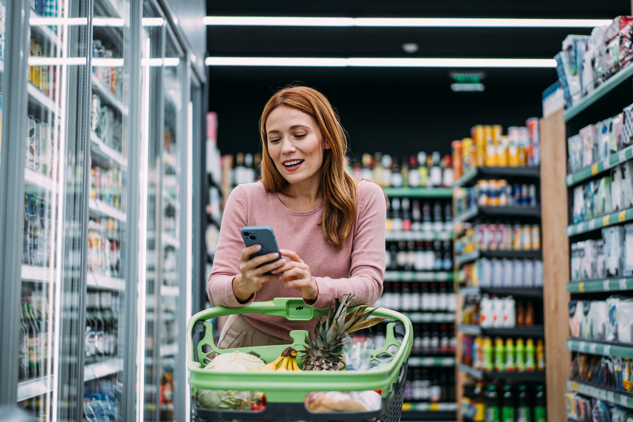 Cheerful woman choosing products and using phone during grocery shopping in supermarket.