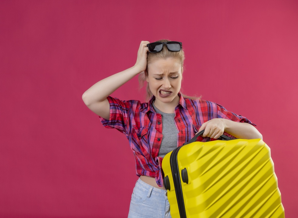sad traveler young girl wearing red shirt in glasses holding suitcase put her hand on head on isolated pink background